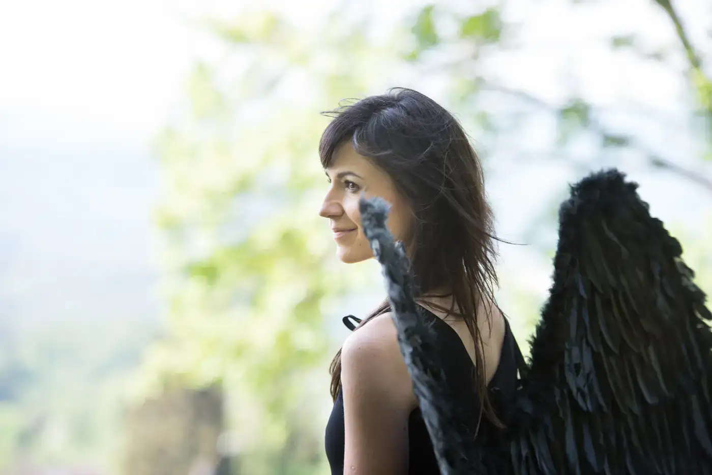 Frau im Freien mit schwarz gefiederten Flügeln, die an einen mystischen Frauenkreis inmitten von grünem Laub erinnern.