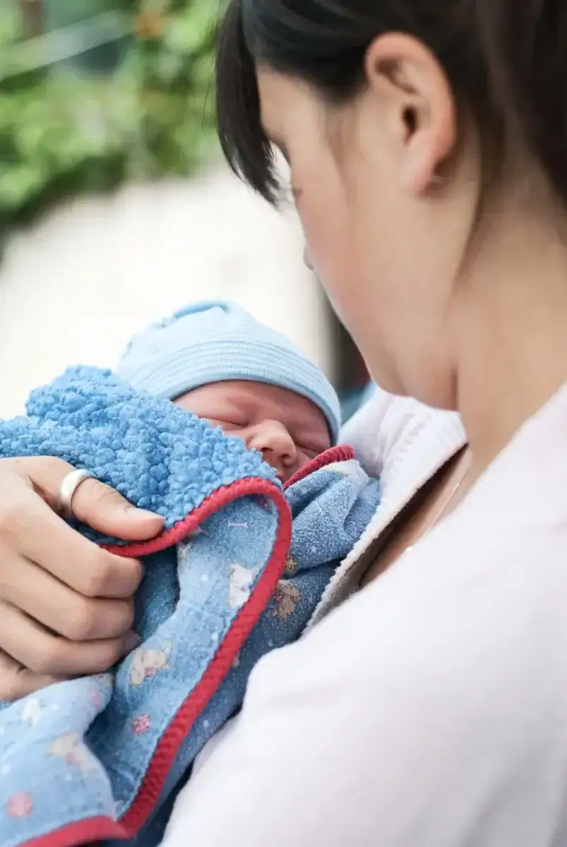 Frau mit neugeborenem Baby, eingewickelt in eine blaue Decke, als Symbol für die Schwangerenbegleitung.