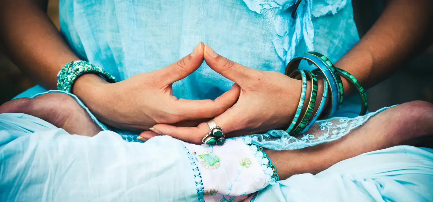 Person in blauer Kleidung meditiert im Schneidersitz, Hände in einer Mudra, trägt Armbänder und Ringe.