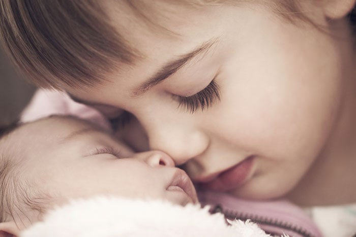 Ein kleines Kind berührt liebevoll die Nasen eines schlafenden Babys, dessen Seele in weiche Decken gehüllt ist.