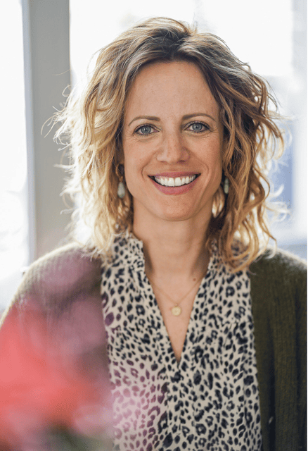 Lächelnde Frau mit gewelltem blondem Haar, die eine Bluse mit Leopardenmuster und eine grüne Strickjacke trägt.