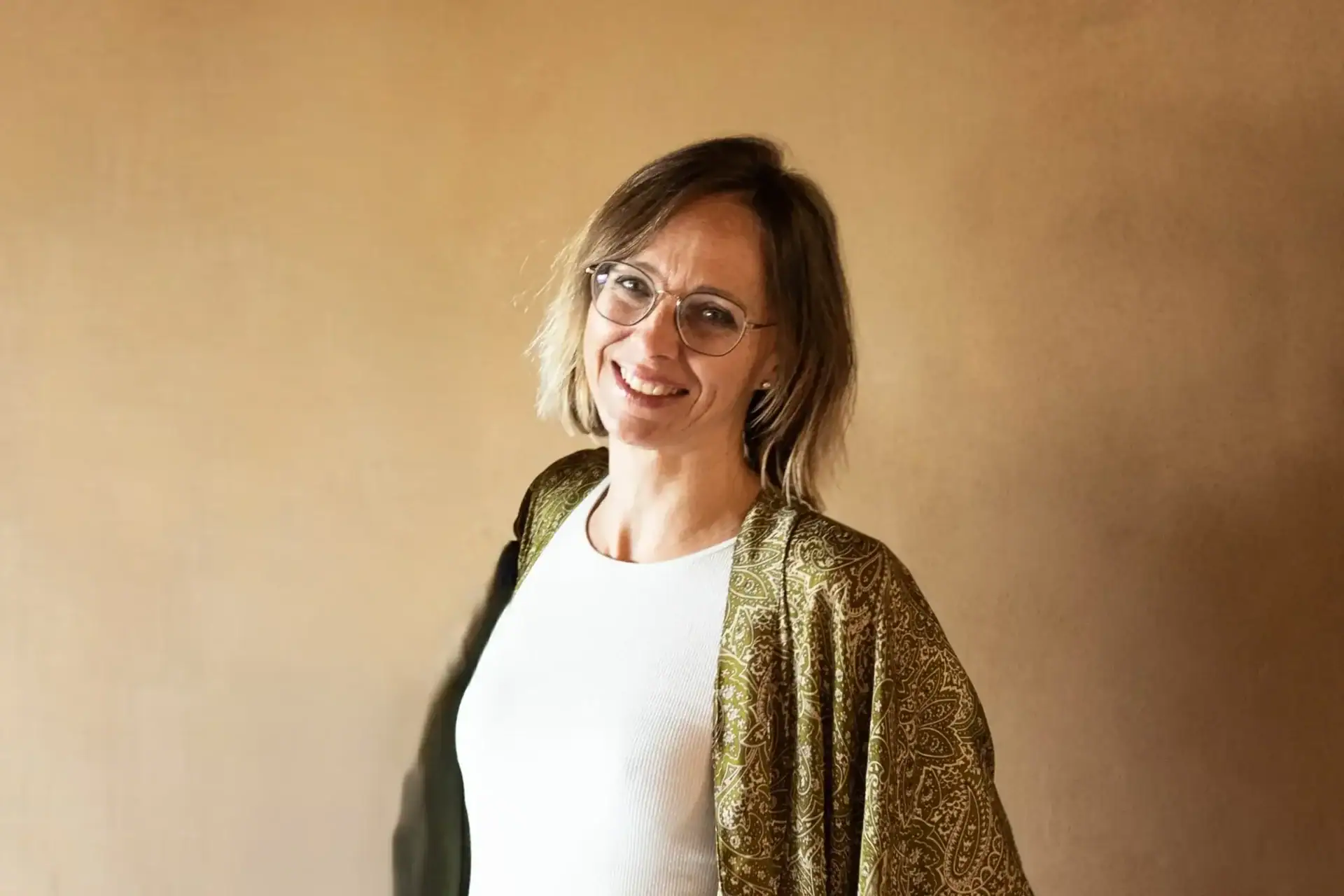 Eine lächelnde Frau mit Brille und gemustertem Schal steht vor einem beigefarbenen Hintergrund und strahlt Ruhe durch Atemarbeit aus.