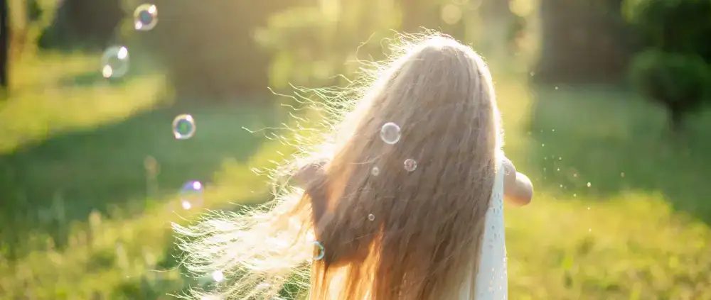 Ein Kind mit langen Haaren genießt die Arbeit mit Kindern, indem es draußen im Sonnenlicht mit schwimmenden Seifenblasen läuft.