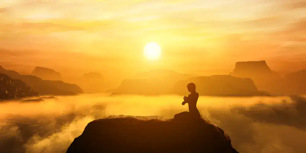 Meditierende Person auf einem Felsen bei Sonnenaufgang, auf der Suche nach Selbsterkenntnis inmitten von nebligen Bergen und goldenem Himmel.