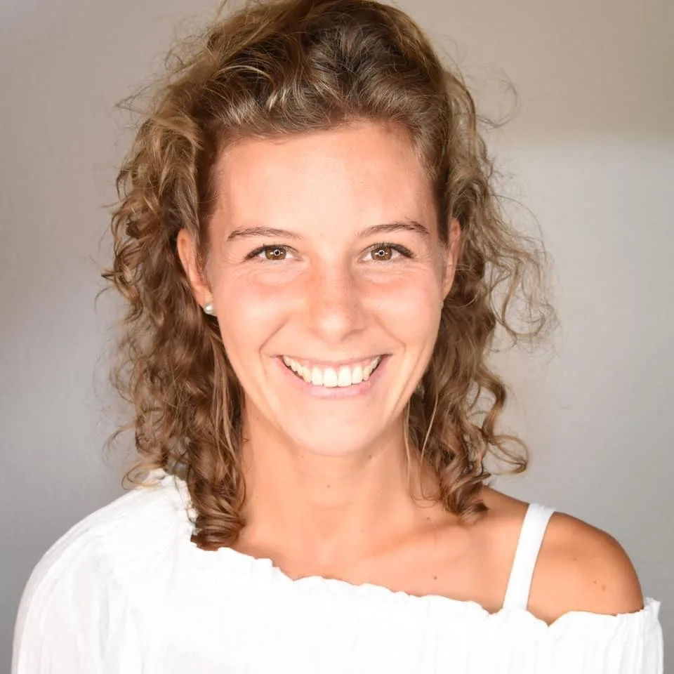 Frau mit lockigem Haar, lächelnd, in einem weißen Oberteil vor einem schlichten Hintergrund.