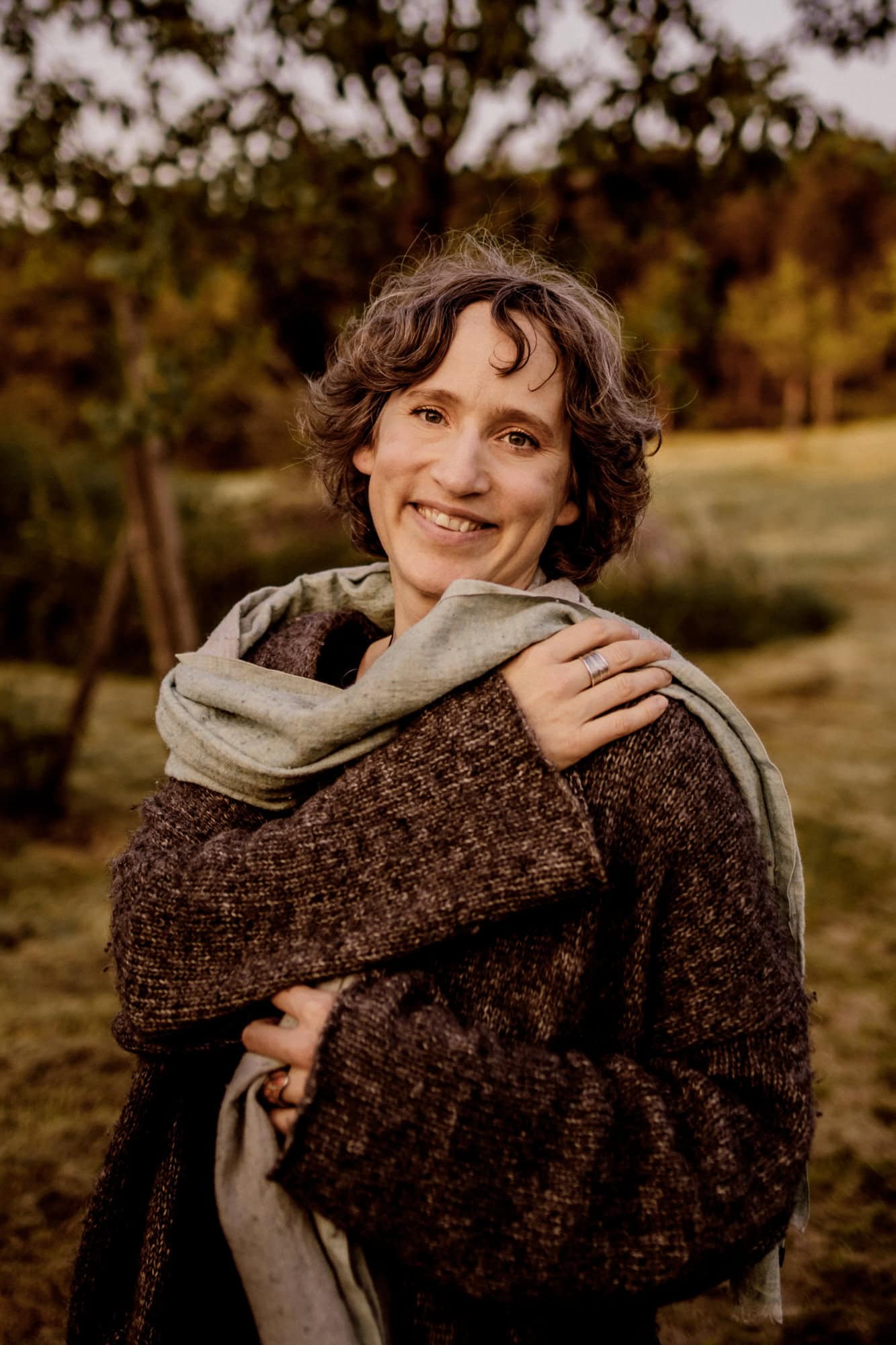 Frau mit kurzen lockigen Haaren in einem braunen Mantel und Schal, lächelnd im Freien auf einem Feld.