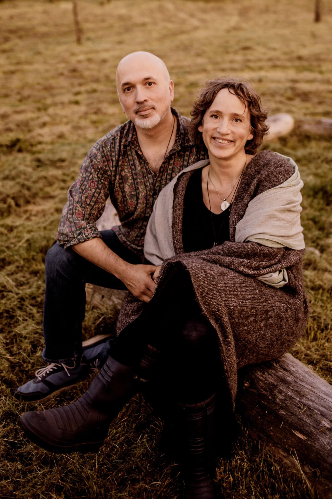 Ein Mann und eine Frau sitzen zusammen auf einem Baumstamm im Freien und lächeln in die Kamera.