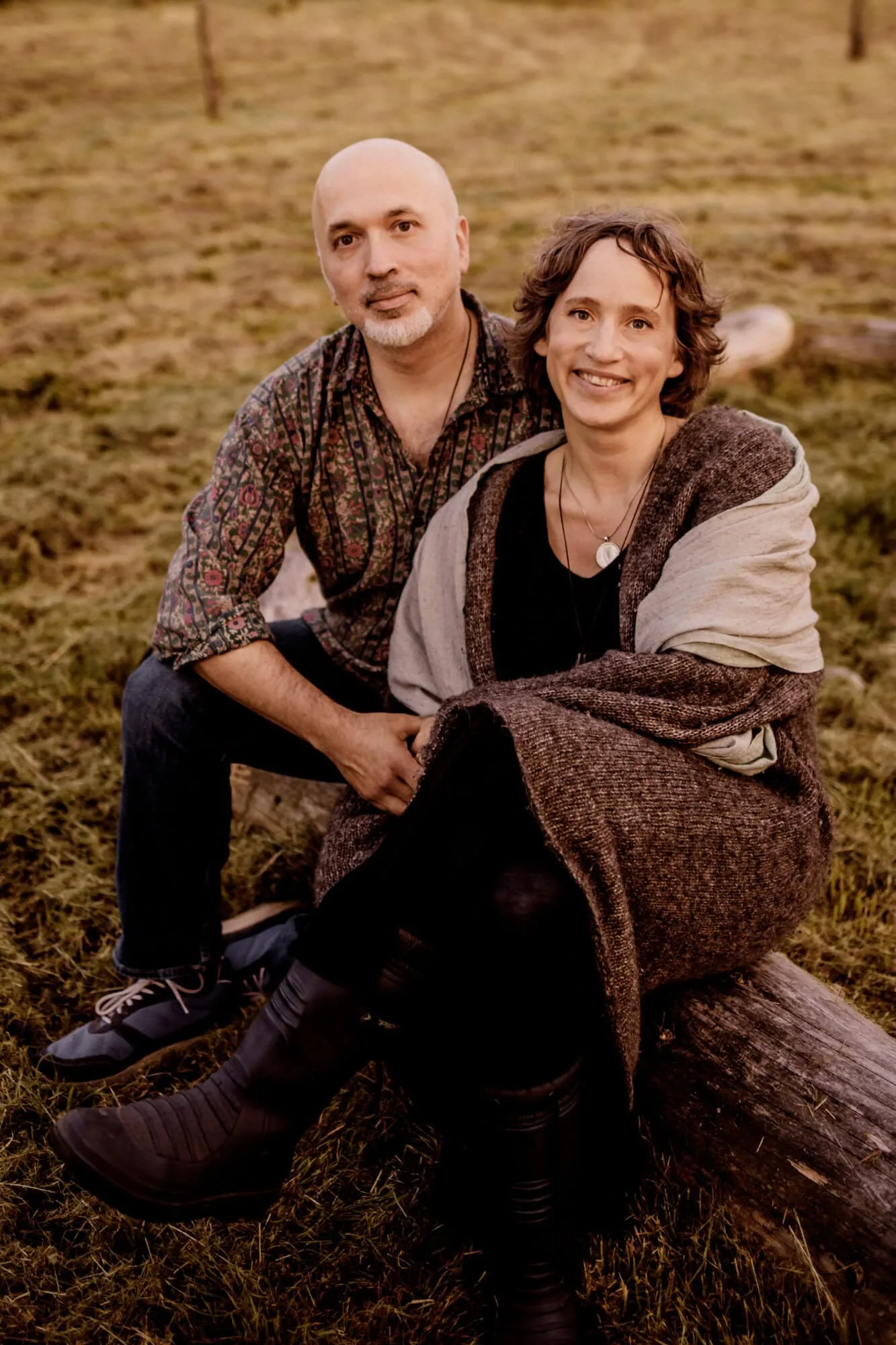 Ein Mann und eine Frau sitzen zusammen auf einem Baumstamm im Freien und lächeln in die Kamera.