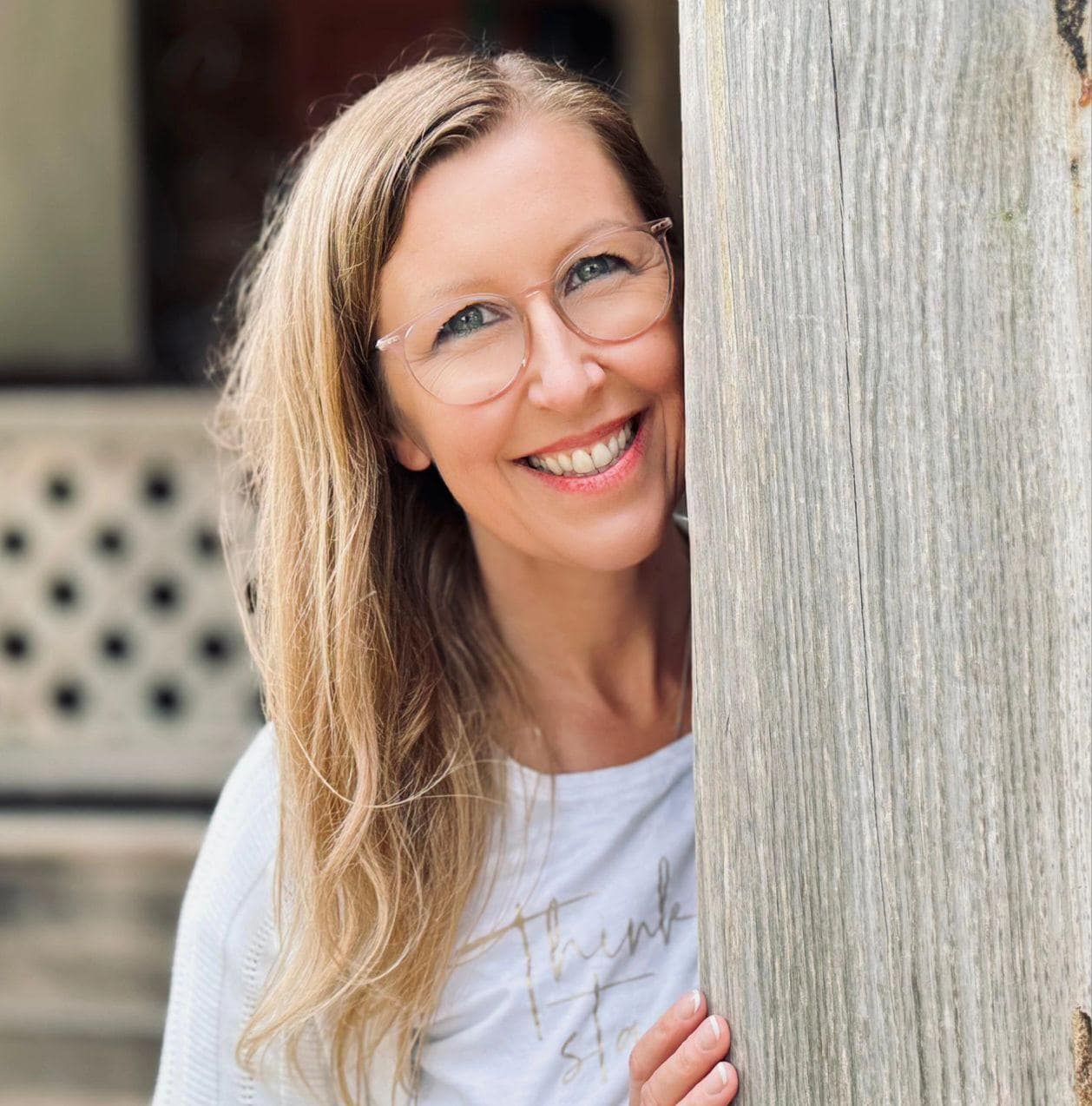 Eine lächelnde Frau mit Brille und langen Haaren lugt hinter einem Holzpfosten hervor.