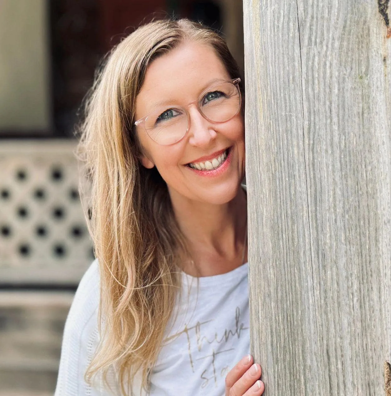 Eine lächelnde Frau mit Brille und langen Haaren lugt hinter einem Holzpfosten hervor.