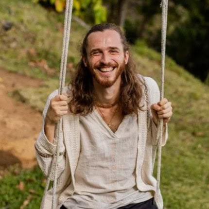 Mann mit langen Haaren, der lächelnd auf einer Schaukel im Freien auf einer Wiese sitzt.