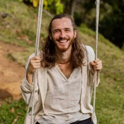 Mann mit langen Haaren, der lächelnd auf einer Schaukel im Freien auf einer Wiese sitzt.