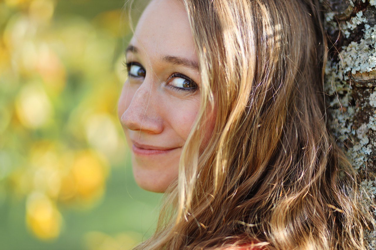 Frau mit langem gewelltem Haar, lächelnd, an einen Baum gelehnt, mit einem unscharfen Hintergrund im Freien.