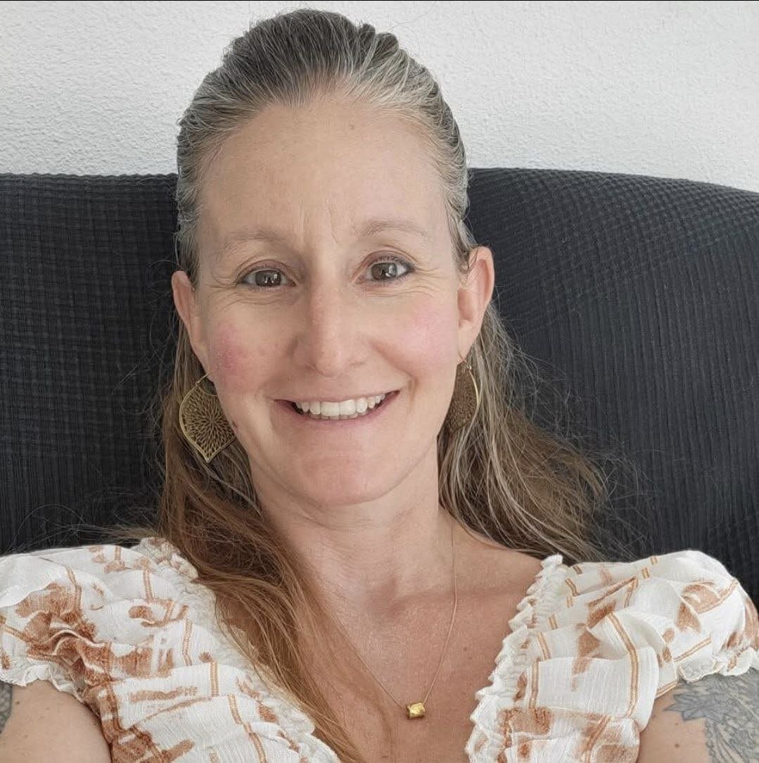 Lächelnde Frau mit langen Haaren, die ein weißes gemustertes Oberteil und goldene Ohrringe trägt, auf einem dunklen Stuhl.