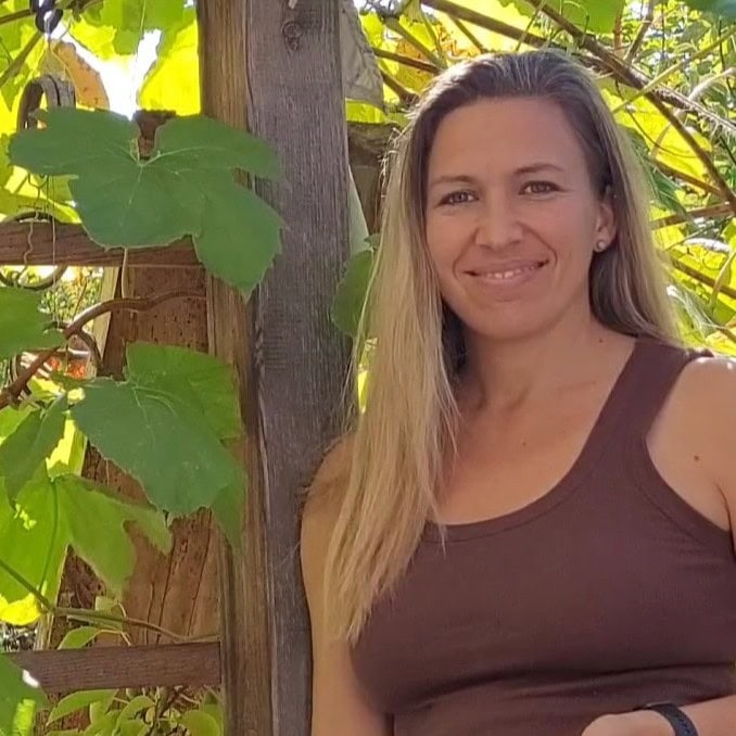 Eine Frau mit langem blondem Haar lächelt an einem Holzspalier mit grünen Blättern.