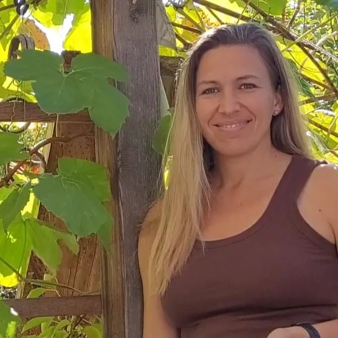 Eine Frau mit langem blondem Haar lächelt an einem Holzspalier mit grünen Blättern.