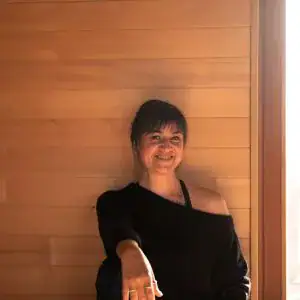 Eine lächelnde Frau mit dunklen Haaren steht vor einer hellen Holzverkleidung und trägt ein schulterfreies schwarzes Oberteil.