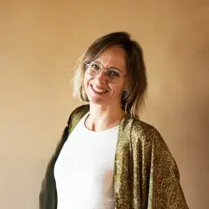 Eine Frau mit kurzem, hellbraunem Haar und Brille lächelt vor einer warmen, beigefarbenen Wand. Sie trägt ein weisses Oberteil und eine gemusterte, grünliche Jacke.